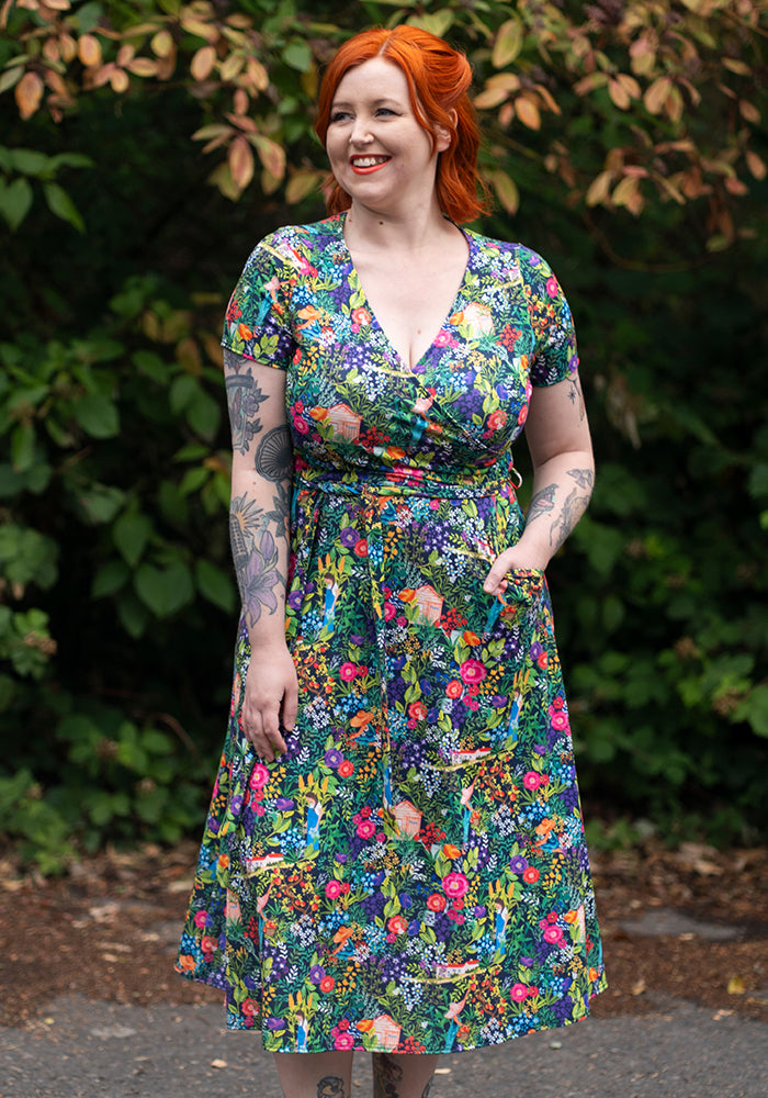 Woman wearing a colourful floral dress standing outdoors with greenery in the background and hand in pocket
