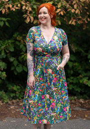 Woman wearing a colourful floral dress standing outdoors with greenery in the background and hand in pocket