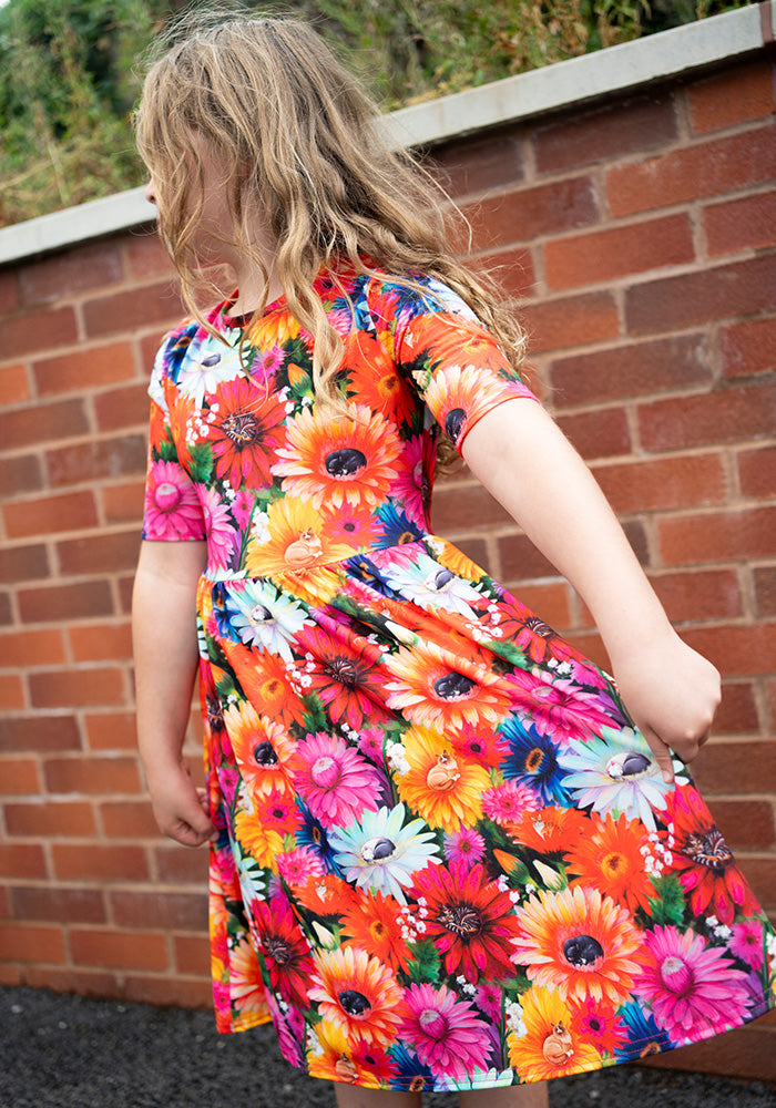 Child wearing a colourful floral and hidden cat print dress in front of a brick wall