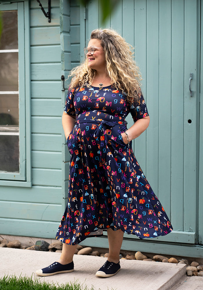 Woman wearing a colourful letters phonics midi dress standing in front of a light blue building.