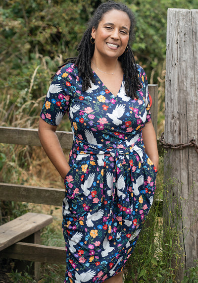 Woman wearing a floral dress with bird pattern standing outdoors near a wooden fence.