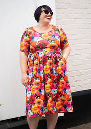 Woman wearing a colourful floral and cat print dress standing against a white brick wall.