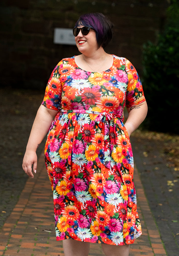 Woman wearing a colourful floral and cat print midi dress with hand in pocket outdoors on a brick path.