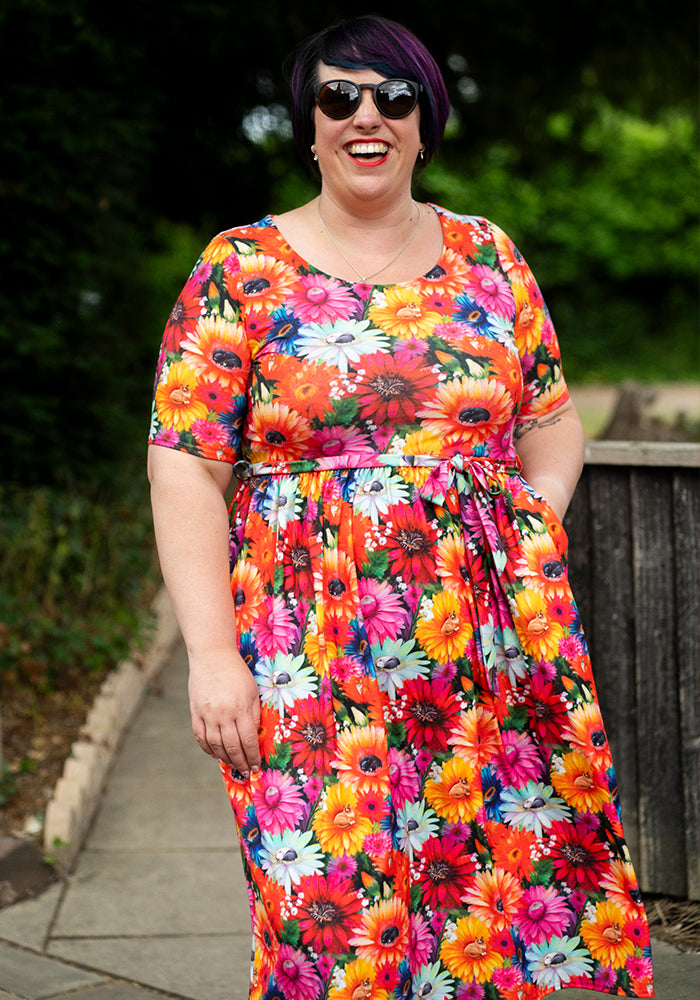 Person wearing a colourful floral and cat print dress outdoors with greenery in the background and hand in pocket