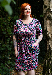 Woman wearing a colourful animal print dress standing next to a tree in a natural setting with hand in pocket